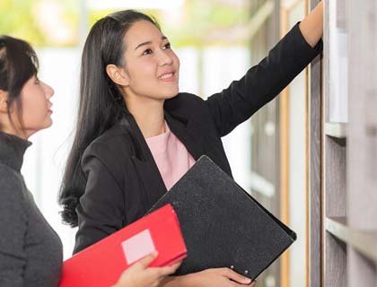 Two women in an office placing files on a shelf.