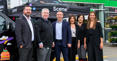 A group of six people stand together smiling in front of a black tent labeled "Scene+" and a background of a building and colorful flowers.
