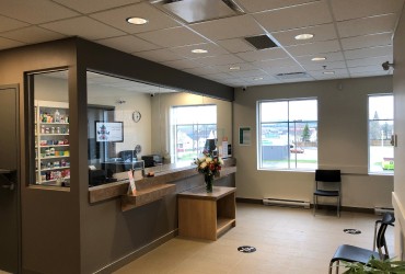 An image of a well-lit reception area with a counter, plexiglass barrier, floral arrangement, and seating.