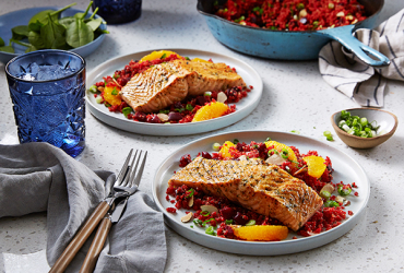 Two plates of grilled salmon with quinoa and orange slices on a white table with blue glasses, utensils, and a pan of more food in the background.