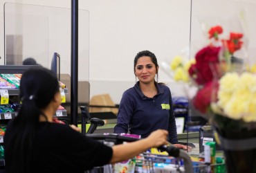 In this image, A cashier in a black shirt attends to a customer at a grocery store checkout.